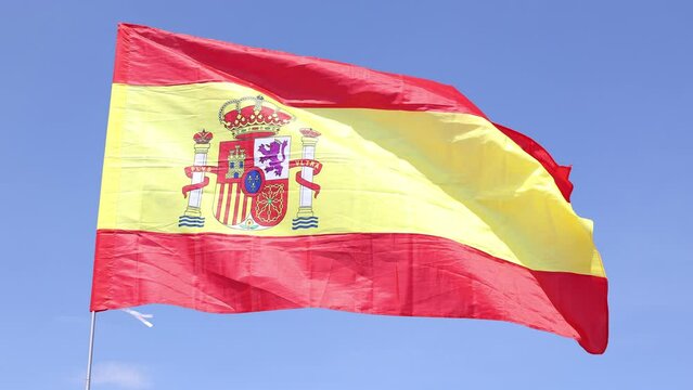 Flag of Spain waving in wind on flagpole against sky with clouds on sunny day.