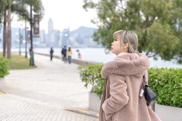 A young Chinese woman in her 20s dressed in winter walking in a park by the sea where you can see the skyscrapers of Kowloon, Hong Kong 香港九龍の高層ビルが見える海沿いの公園を散歩する冬の格好をした20代の若い中国人女性