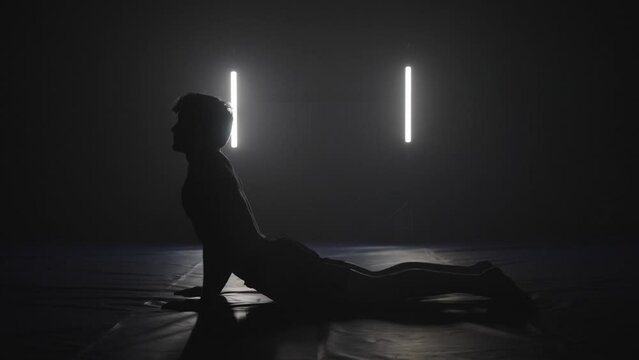 young man doing sphinx yoga pose in dark studio, silhouette yoga pose
