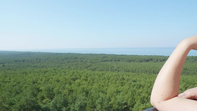 A woman admires the panoramic view of the lush green forest and the sea from the top of the Stilo lighthouse in Stilo, Poland ,while birds fly around.