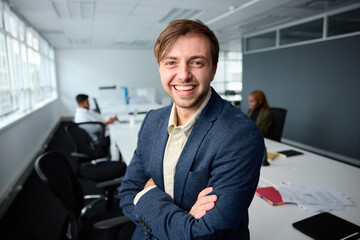 Fototapeta na wymiar Happy young businessman in businesswear with arms crossed by desk with coworkers in office