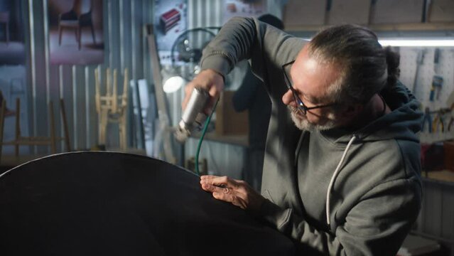 Mature carpenter makes chair upholstery using staple gun in modern workshop for making and renovation furniture. Another specialist assembles shelf in the background. Handmade and entrepreneurship.