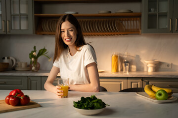A young woman is seated at a kitchen counter, sipping from a glass of fresh juice, with a soft...