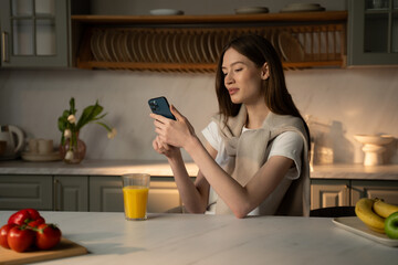 A young woman is casually seated at a kitchen counter, engrossed in her smartphone. She is...