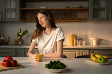 A young woman is seated at a kitchen counter, sipping from a glass of fresh juice, with a soft...