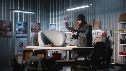 Male carpenter or designer takes picture of chair using tablet computer working in modern workshop for making and renovation furniture. Craftsman assembles shelf in the background. Handmade concept.
