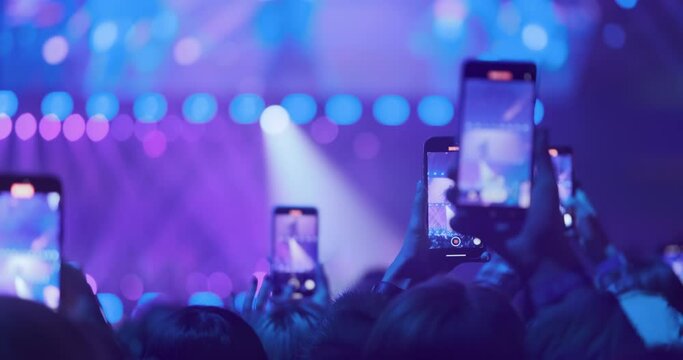 Close up, many phone screens capture bright blue violet concert scene in spotlight. Spectators group together capturing happy moments on phones. Smartphone video recording, filming of important events