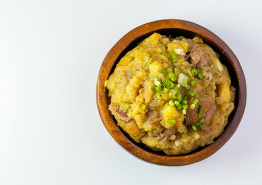 Mashed plantains with garlic and pork in a wooden bowl garnished with green onions, top view isolated on white background. Caribbean mofongo dish concept. Design for menu and culinary blog