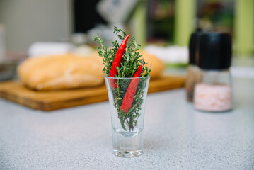 Fresh Thyme and Red Chili Pepper in Glass. A sprig of fresh thyme paired with a vibrant red chili pepper in a clear glass shot, set against a kitchen backdrop.