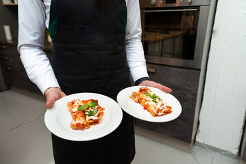 Waiter Serving Plates of Gourmet Lasagna. A waiter in a black apron presents two plates of freshly baked lasagna garnished with basil, ready to be served.
