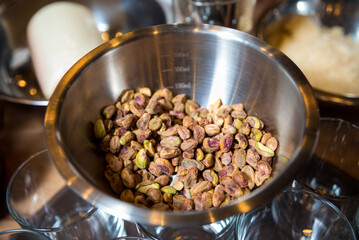 Pistachio Nuts in Stainless Steel Mixing Bowl. A bowl of shelled pistachios ready for cooking or baking, with kitchen utensils and ingredients in the background.