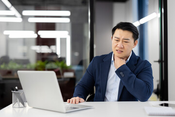 Sick Asian young male office worker working on a notebook, sitting at a table, holding his hand to his neck, feeling a strong pain in his throat.