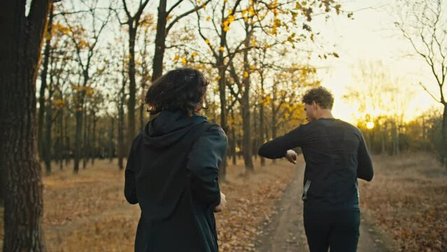 Rear view of two brunette guys in black sportswear running along an earthen path with fallen autumn leaves in the morning at Sunrise