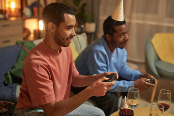 Side view portrait of two adult friends playing video game together sitting on couch at home and holding controllers copy space