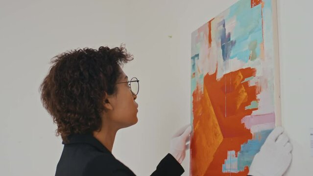 Close-up shot of young African American female art museum employee in white gloves carefully hanging up bright modernist style masterpiece on wall, while making preparations for exhibition opening