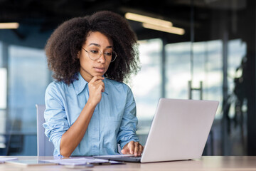 A serious young African American woman is sitting in the office, at desk in a blue shirt and is...