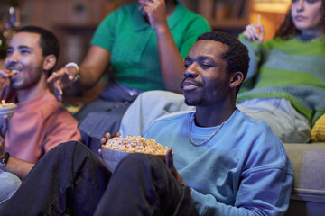 Portrait of Black young man watching TV in dark with group of friends and holding bowl of popcorn