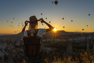 Mesmerizing shot of a young woman in a hat with a craft backpack overlooking balloons in the rays...