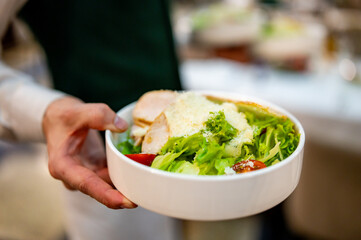 Close-up of a person in a white shirt holding a bowl of fresh Caesar salad with lettuce, tomatoes, cheese, and grilled chicken in an indoor setting.