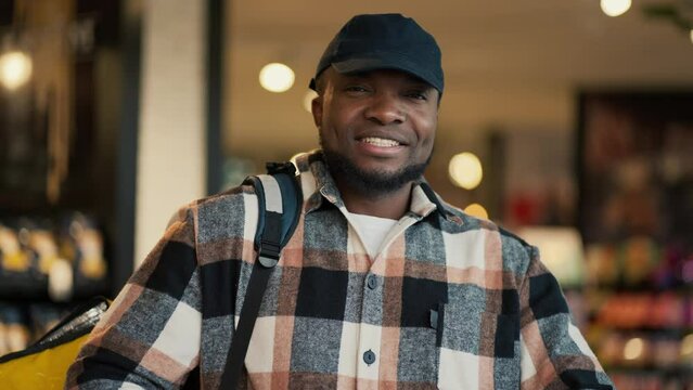 Portrait of a happy man with Black skin in a checkered shirt and a black cap who works as a delivery man and carries a large yellow bag on his shoulders while looking for the necessary products in a