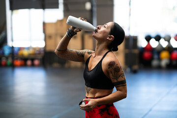 Tired strong mature sportswoman drinking water from a flask indoors