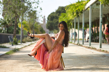 Young woman, Hispanic, beautiful, brunette, with an elegant salmon-colored dress, posing sensual and provocative, sitting on a chair outdoors. Concept of beauty, fashion, trend, elegance.