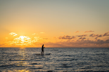 Black sunset silhouette of paddle boarder standing on SUP. Lone paddle board surfer man is surfing...