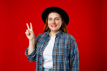 Photo of happy woman in hat makes peace gesture, 30s girl shows v sign on red studio background. Emotional, young face. - Powered by Adobe