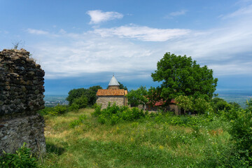 Fototapeta na wymiar Dome of old stone church with orange tiled roof. Stone wall and green grass around. Bright blue sky with clouds. Alazani valley on background.