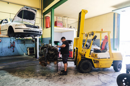 Auto mechanic dismantling engine parts near forklift at workshop