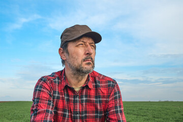 Portrait of mid-adult male farmer wearing brown cap and red plaid shirt in agricultural field, looking into distance, thinking and planning