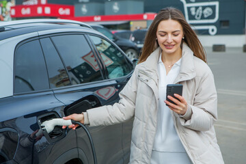 Young beautiful woman traveling by electric car having stop at charging station. Electric car...