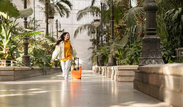 Happy woman walking with suitcase near plants