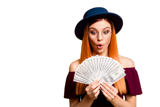 Portrait of red straight-haired attractive beautiful nice amazed young girl, wearing spectacles and dark blue hat, showing fan of hundreds dollars cash. Isolated over bright vivid yellow background