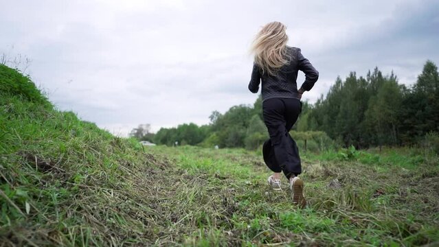 A trim woman with blonde hair runs away against the background of a field. The concept of running and country life.