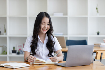 Happy young asian woman wearing earphones and using laptop computer at desk in office, Female asian student with laptop computer, College female student learning online.