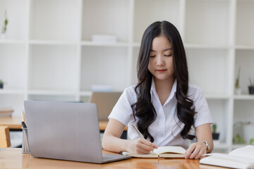 Happy young asian woman wearing earphones and using laptop computer at desk in office, Female asian student with laptop computer, College female student learning online.