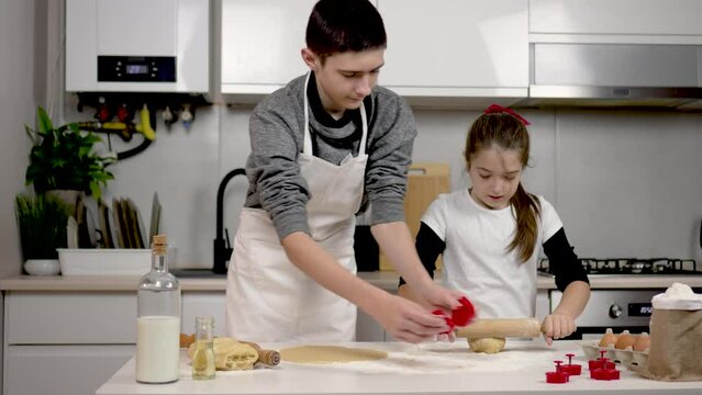 Girl rolls dough with rolling pin on table. Boy makes cookies with molds by cutting out figures at kitchen. Teenage children brother and sister cooking together. Happy family kids dream lifestyle