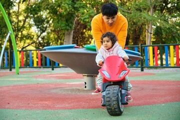 Latin father playing with his one year old daughter on a balance bike in a park.