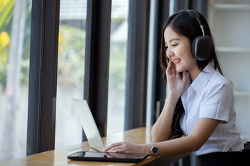 Happy young asian woman wearing earphones and using laptop computer at desk in office, Female asian student with laptop computer, College female student learning online.