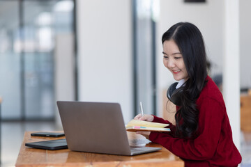 Happy young asian woman wearing earphones and using laptop computer at desk in office, Female student with stack of books and laptop.