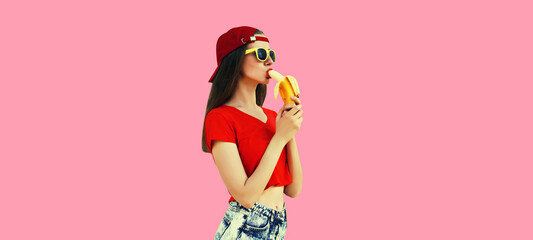 Stylish young woman eating banana in red baseball cap on pink studio background