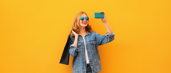 Happy smiling young woman taking selfie with phone holding shopping bags on yellow background