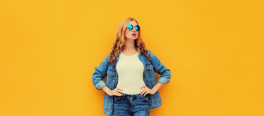 Beautiful modern young blonde woman posing in denim jacket, round sunglasses on yellow background
