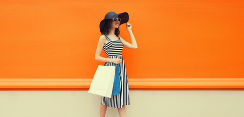 Beautiful happy woman model with shopping bags in summer black hat, dress on orange background