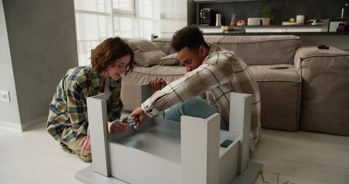 Confident and happy young man with Black skin color in a beige shirt is repairing and assembling a table together with his young adult girlfriend with a bob hairstyle in a plaid shirt sitting on the