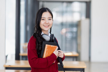 Fototapeta premium Happy young asian woman wearing earphones and holding a book in a classroom at high school.