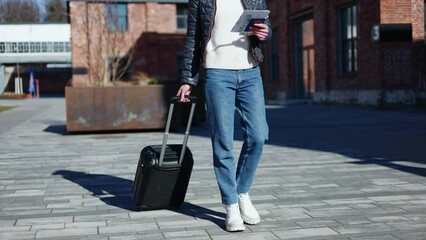 Cropped view of unknown female walking with travel suitcase and holding smartphone with document package. Relaxed woman wearing jeans and sweater and going spending weekend in all inclusive resort.