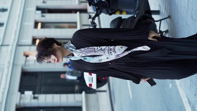 Vertical video. An Asian young woman walks down the street in a black long dress with a fashionable silk scarf around her neck against the backdrop of a business center during the daytime. slow motion