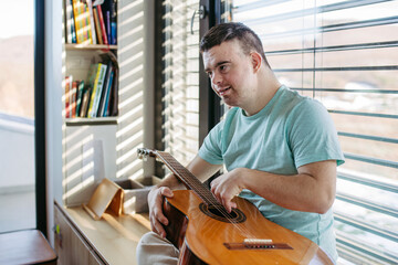 Young man with down syndrome playing acoustic guitar, sitting by window, holding and strumming...